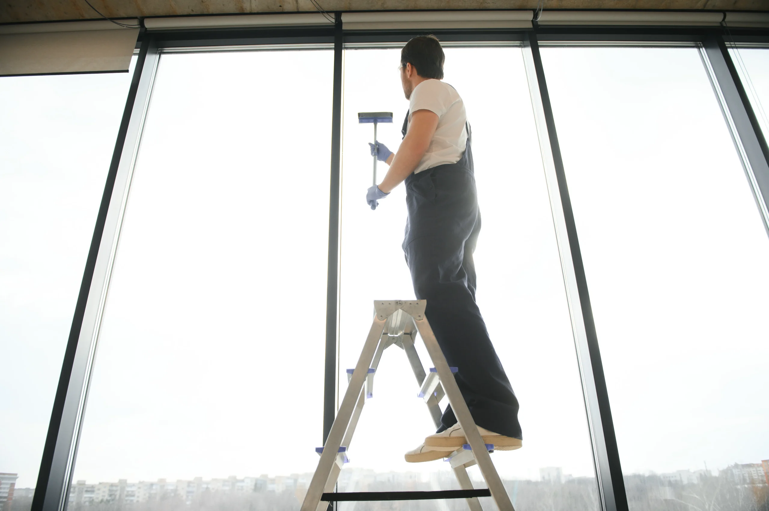 Professional cleaner using a stable ladder during window cleaning of tall ground-floor windows with high ceilings.